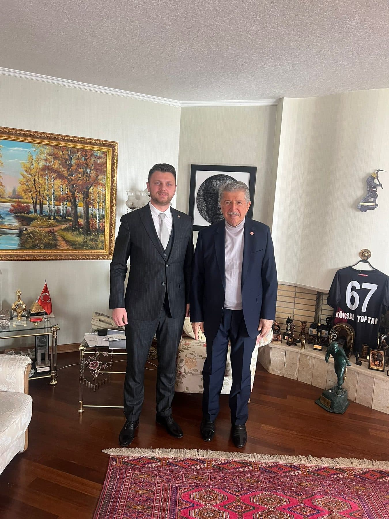 Ömer Yazıcıoğlu with his host in a home displaying a landscape painting and a named sports jersey